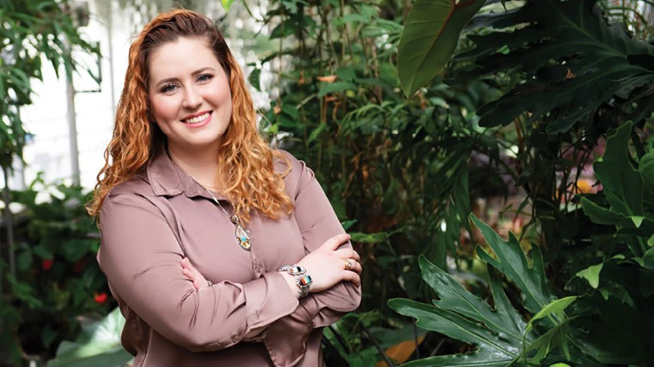 A woman in a mauve blouse smiles at the camera with her arms crossed. She stands in front of a number of large green plants.
