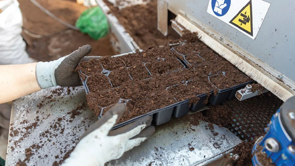 Two pairs of hands with gloves on load a black tray full of soil into a machine.