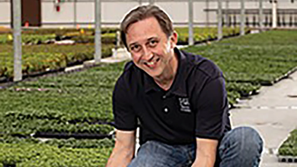 A smiling man wearing a black polo shirt and blue jeans kneels in a greenhouse surrounded by green plants.