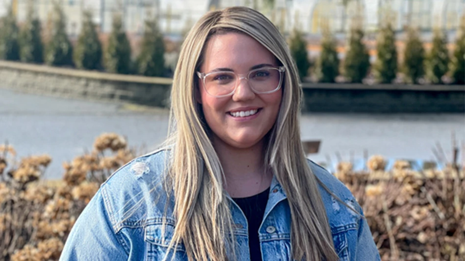 A smiling woman with long blonde hair and glasses wearing a denim jacket and black shirt underneath stands in front of a scenic background with trees, plants, water and a greenhouse.