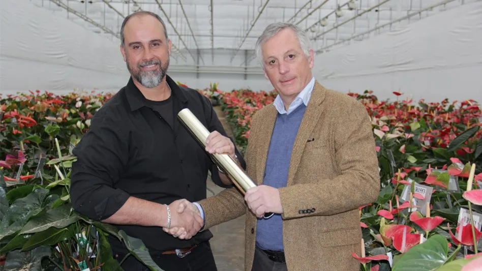 Two men are pictured in a greenhouse with flowers in the background. They are shaking each other's hands and also holding onto a white baton. The man on the left has short dark hair and a grayish-black beard and wears a black dress shirt. The man on the right has short gray hair and wears a blue dress shirt, blue sweater and a tan suit jacket.