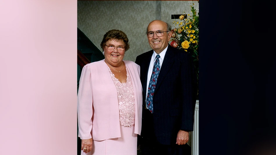 A smiling man and woman pose for a photo. The woman has short brown hair and wears glasses and a pink top, jacket and skirt. The man is mostly bald with some gray hear and wears glasses, a black suit, a white dress shirt and a blue and red tie.