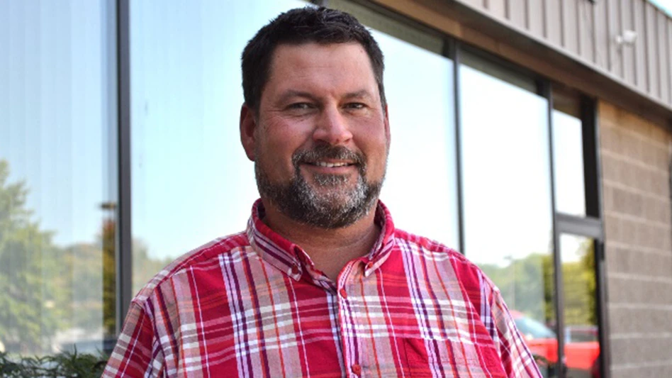 A smiling man with short dark brown hair and a short dark brown and gray beard stands in front of a brick building with large windows. He wears a red and white plaid button-up shirt.