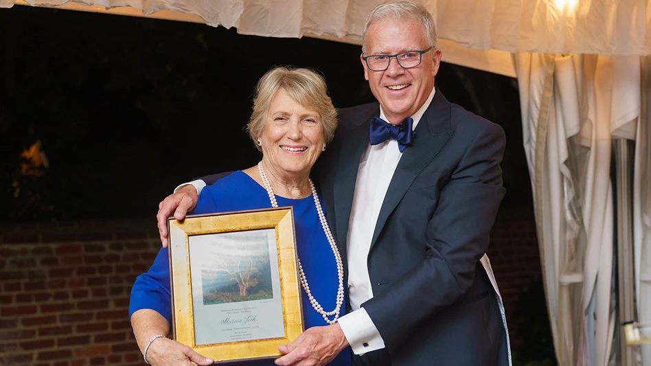 A smiling man with short white hair and glasses wearing a navy blue suit jacket, white dress shirt and navy blue bow ties stands with his arms around a smiling woman with short blonde hair wearing a blue top and long pearl necklace. They hold a gold-framed plaque.