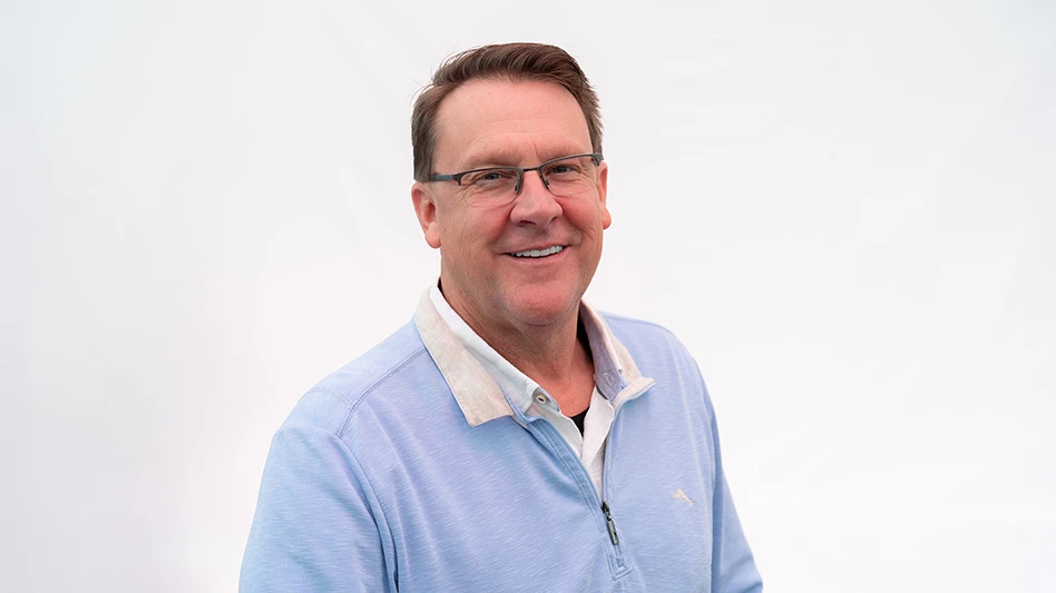 A smiling man with short brown hair wearing a light blue collared shirt and glasses.