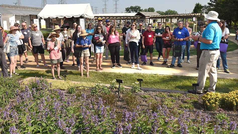 A group of people listen to a man in a blue shirt, tan pants and tan hat speak. They're standing in front of a garden full of purple flowers.