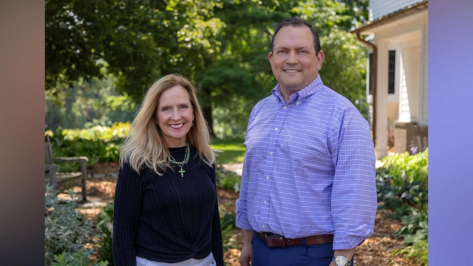 A woman in a black sweater and a man in a blue shirt standing in front of a plant-filled landscape.