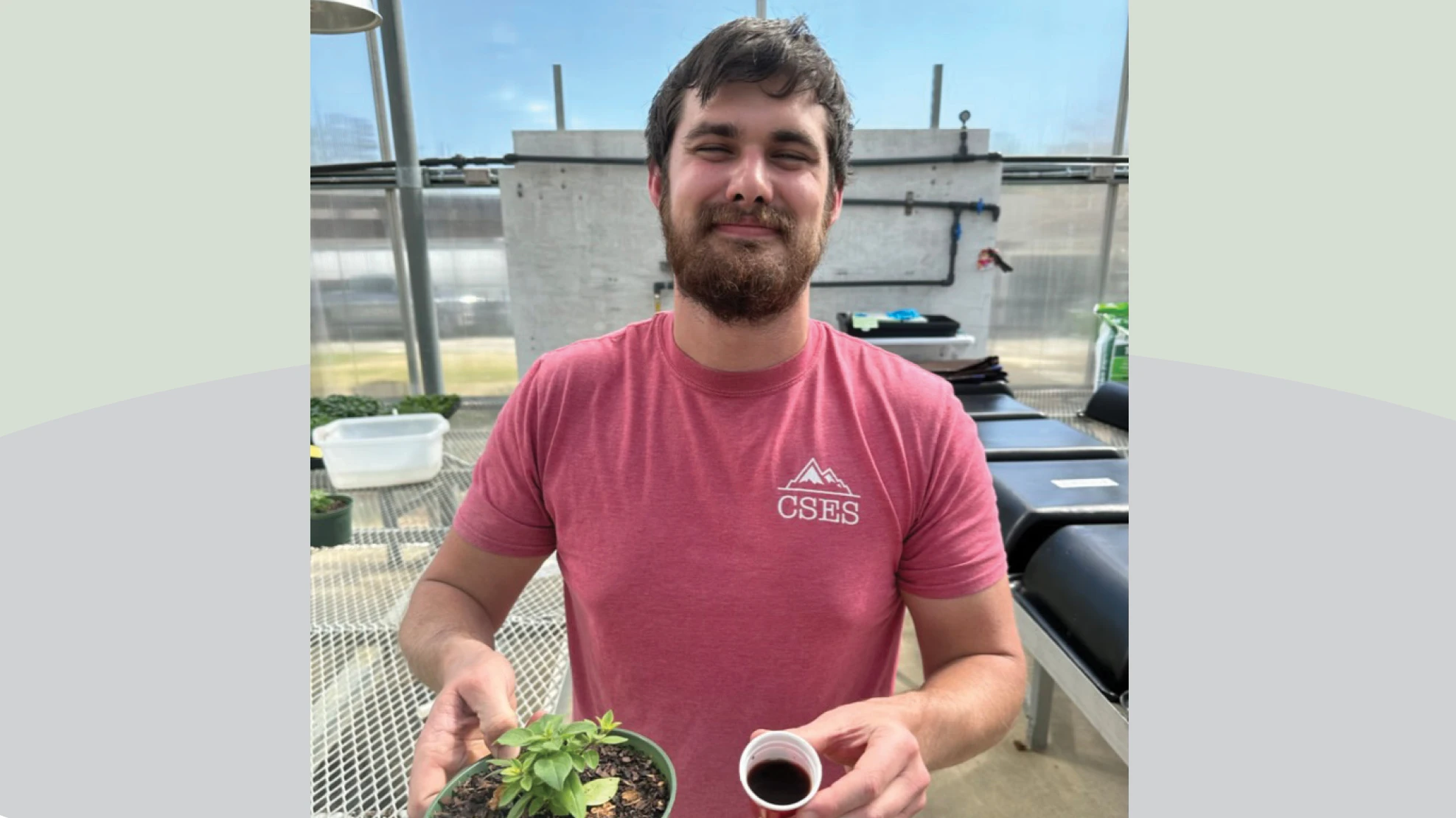 A smiling man with brown hair and facial hair wearing a red T-shirt, standing in a greenhouse and holding a potted green plant and one hand and a small plastic cup of brown liquid in the other hand.