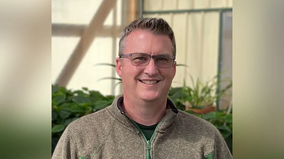 A headshot photo of a smiling man with short brown hair wearing glasses and a brown pullover sweater standing in a greenhouse.