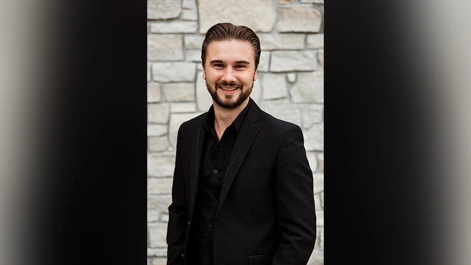 A smiling man with brown hair and a short brown beard wearing a black shirt and black jacket and standing in front of a gray stone wall.