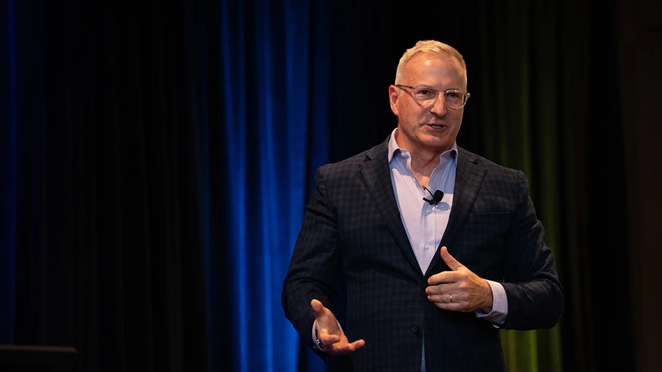 A man with short white hear wearing clear glasses, a navy blue checked suit jacket and a light blue dress shirt gestures with both hands while he speaks on a stage.