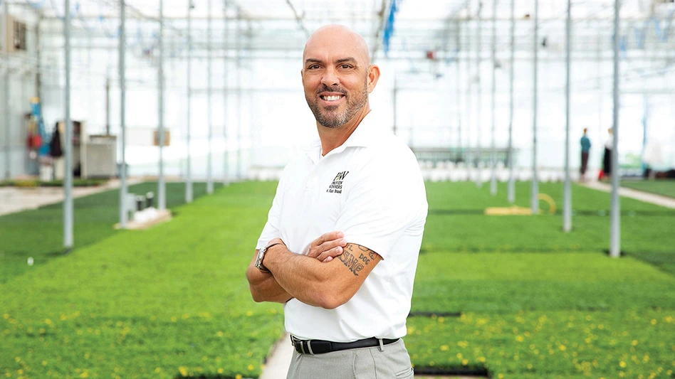 A smiling bald man wearing a white polo shirt poses for a photo with his arms crossed while standing in front of a greenhouse full of green plants.