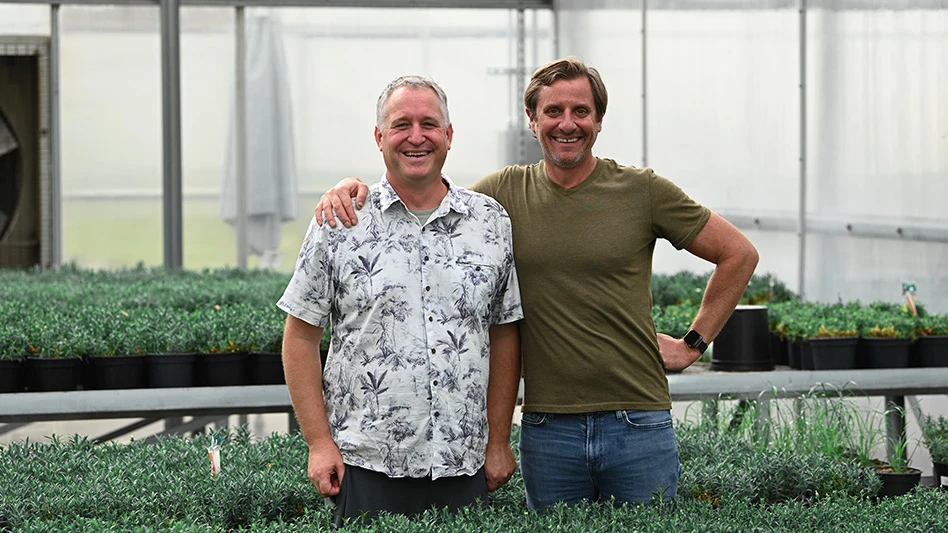 Two smiling men, one with his arm around the other, pose for a photo in a greenhouse surrounded by small green plants on benches.
