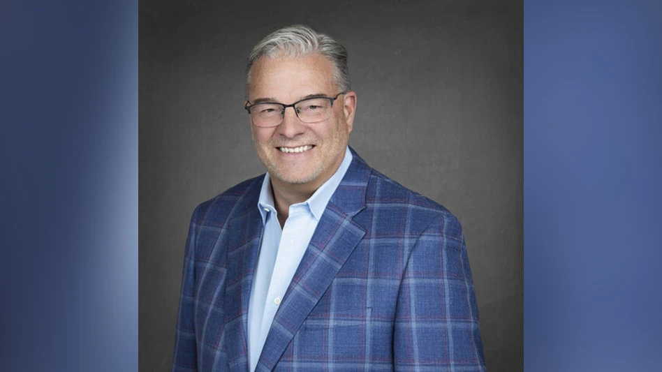 A headshot photo of a smiling man in front of a gray background. He has short gray hair and is wearing glasses, a light blue dress shirt and a dark blue checked sport coat.