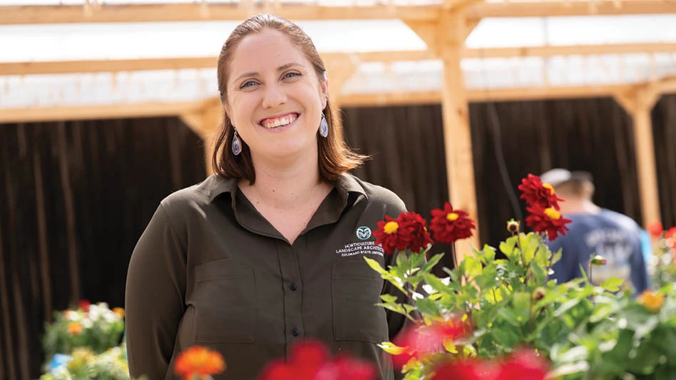 A smiling woman with brown shoulder-length hair wearing an olive green button-up shirt and standing amid flowers and a wooden pergola.