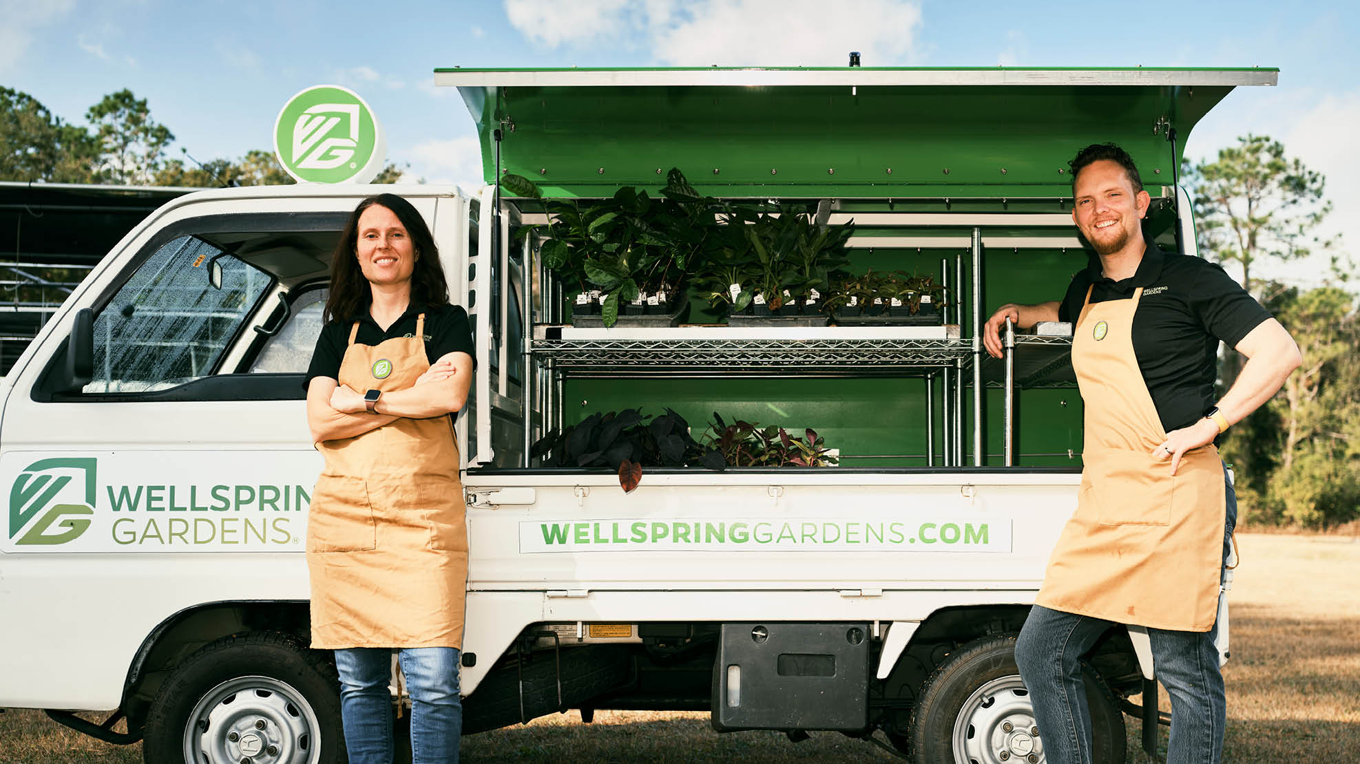 A smiling man and woman, both wearing black shirts and tan aprons, lean against a white and green truck with green plants in the bed.