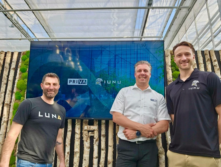 Three smiling men pose in front of a blue screen in a greenhouse.