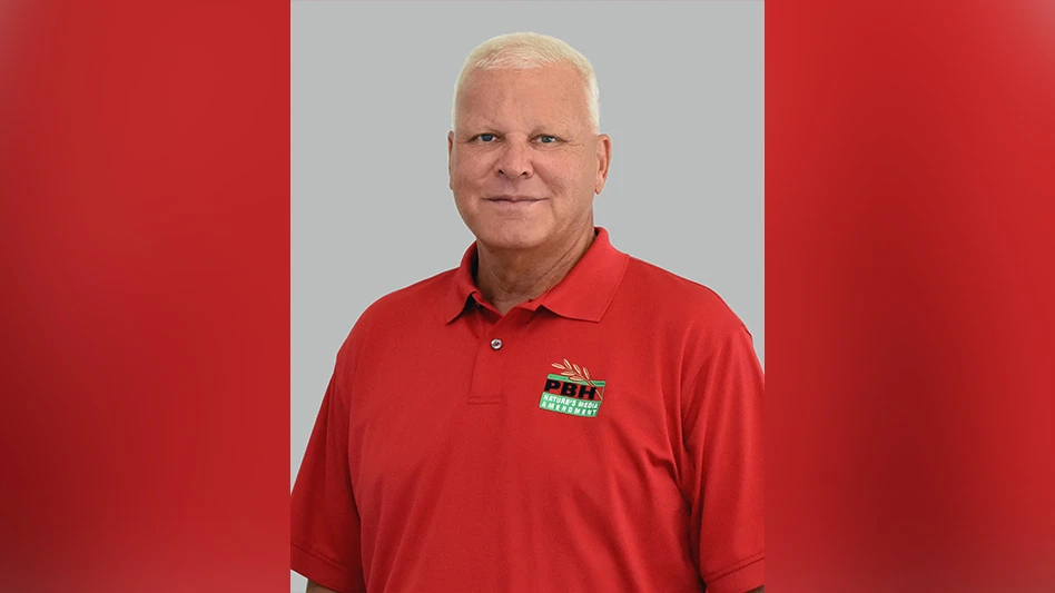 A man with short white hear wearing a red polo shirt and standing in front of a gray background.