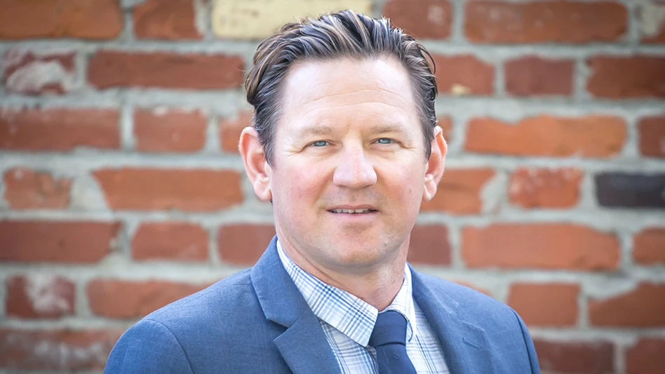 A headshot photo of a man with short brown hair wearing a blue suit jacket, white and blue shirt and blue tie. He's standing in front of a red brick wall.