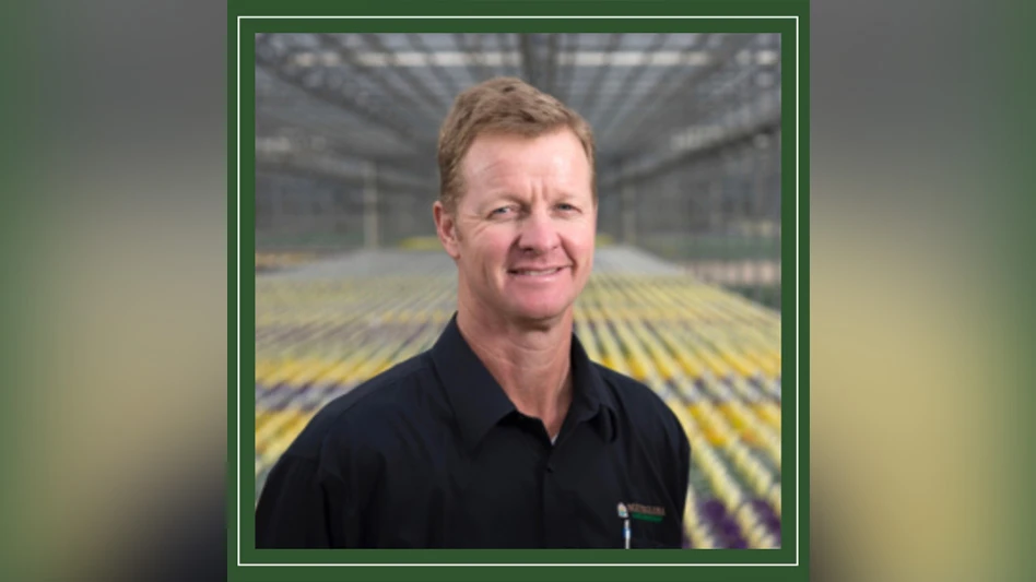 A headshot photo of a smiling man with short red-brown hair wearing a black shirt and standing in a greenhouse.