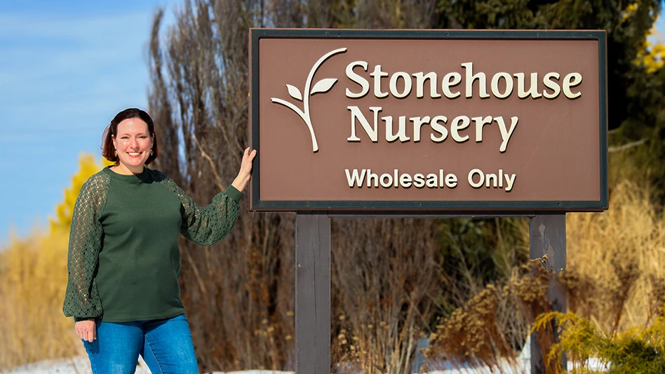 A smiling woman with short brown hair and wearing a green long-sleeved shirt and blue jeans stands next to a brown, rectangular sign on two brown posts reading Stonehouse Nursery Wholesale Only in white letters. She's holding the sign with one hand, and trees and shrubs are visible in the winter background.