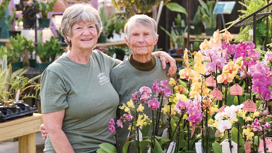 Two smiling women with their arms around each other pose in front of a table full of potted pink and yellow orchids.