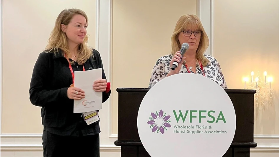 One woman holding a microphone stands behind a brown podium with a white circular sign. A second woman holding white papers is to the left of her.