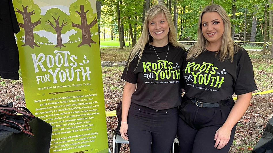 Two smiling women with long blonde hair and wearing black T-shirts with green and white text reading Roots for Youth and black jeans. They stand next to a green sign with white text reading Roots for Youth and graphics of trees.